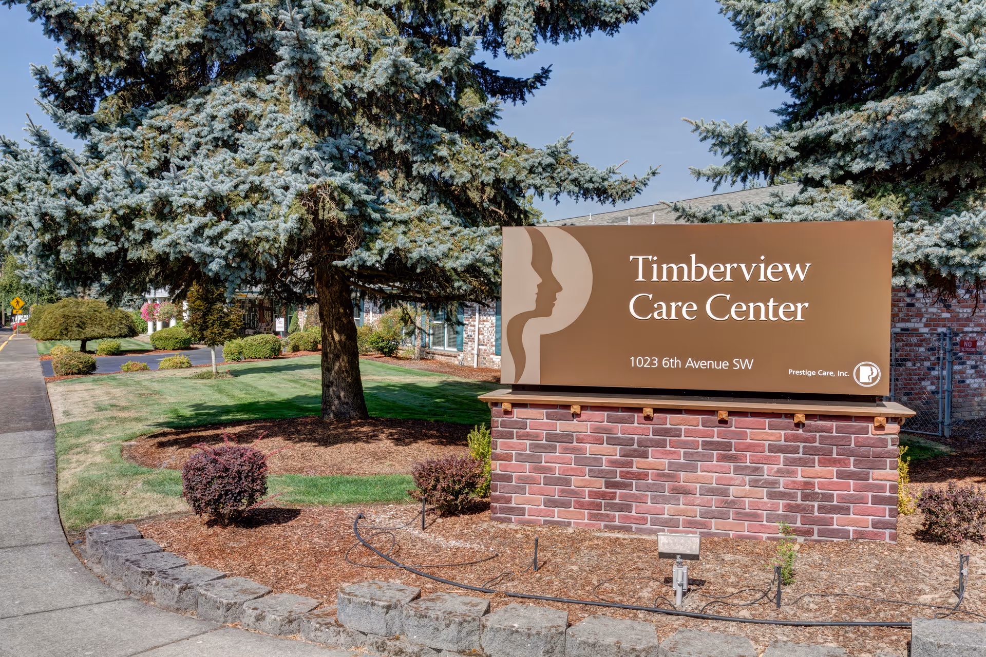 Outdoor view of a landscaped area with a large evergreen tree and a brick sign that reads 'Timberview Care Center, 1023 6th Avenue SW, Prestige Care, Inc.'