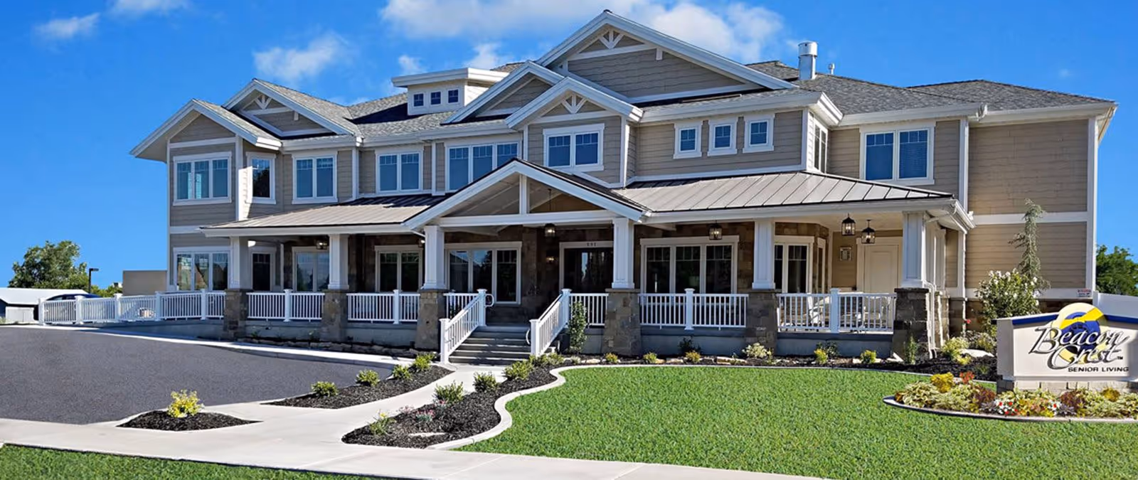 Front exterior view of a large two-story senior living facility building with beige siding, multiple windows, a covered porch with white railings, and a landscaped lawn with flower beds. A sign in front reads 'Beacon Crest Senior Living'.