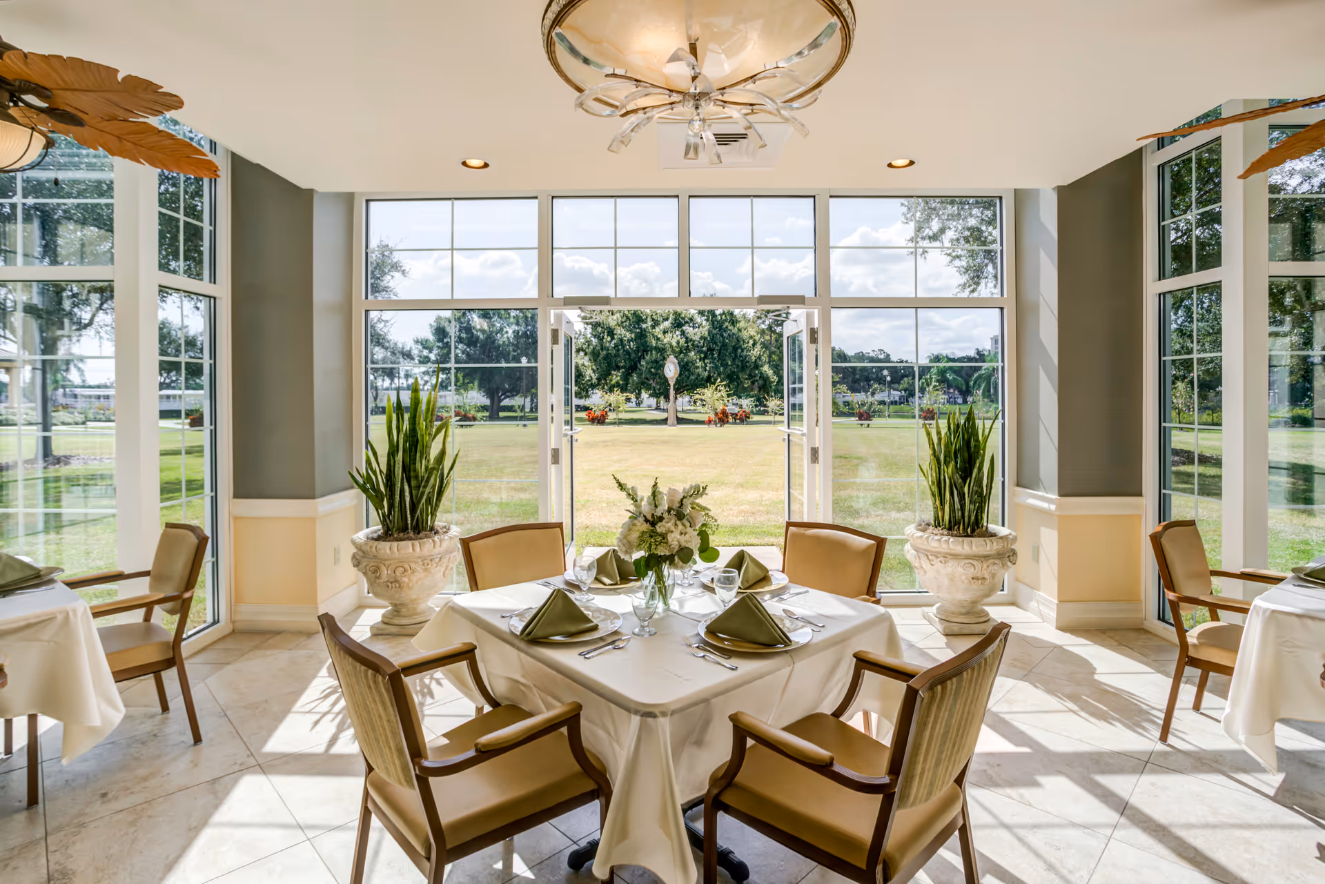 Bright dining room with a round table set for six, large windows, and greenery outside.