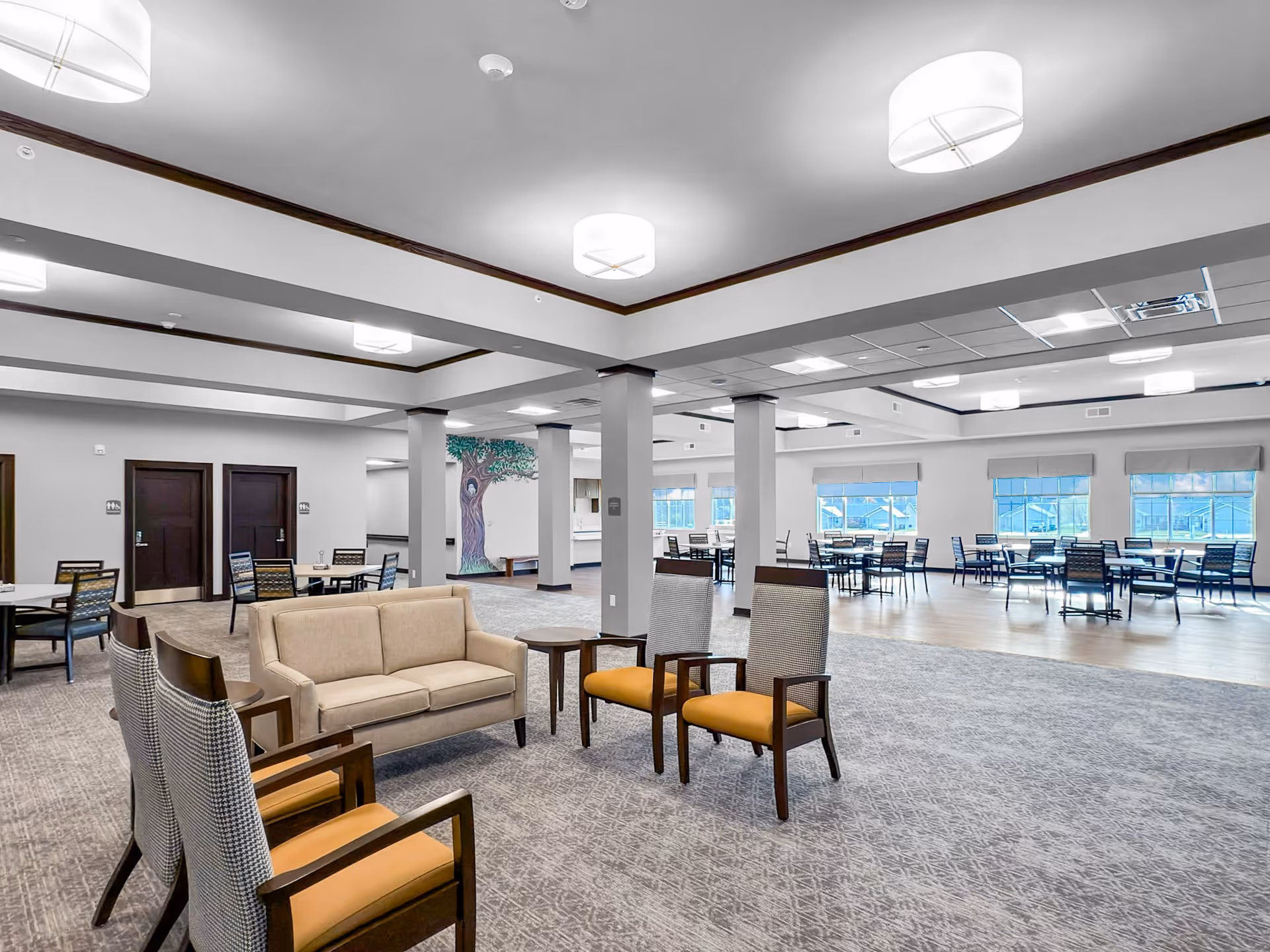 Spacious senior living community dining and lounge area with seating clusters, tables, and large windows.