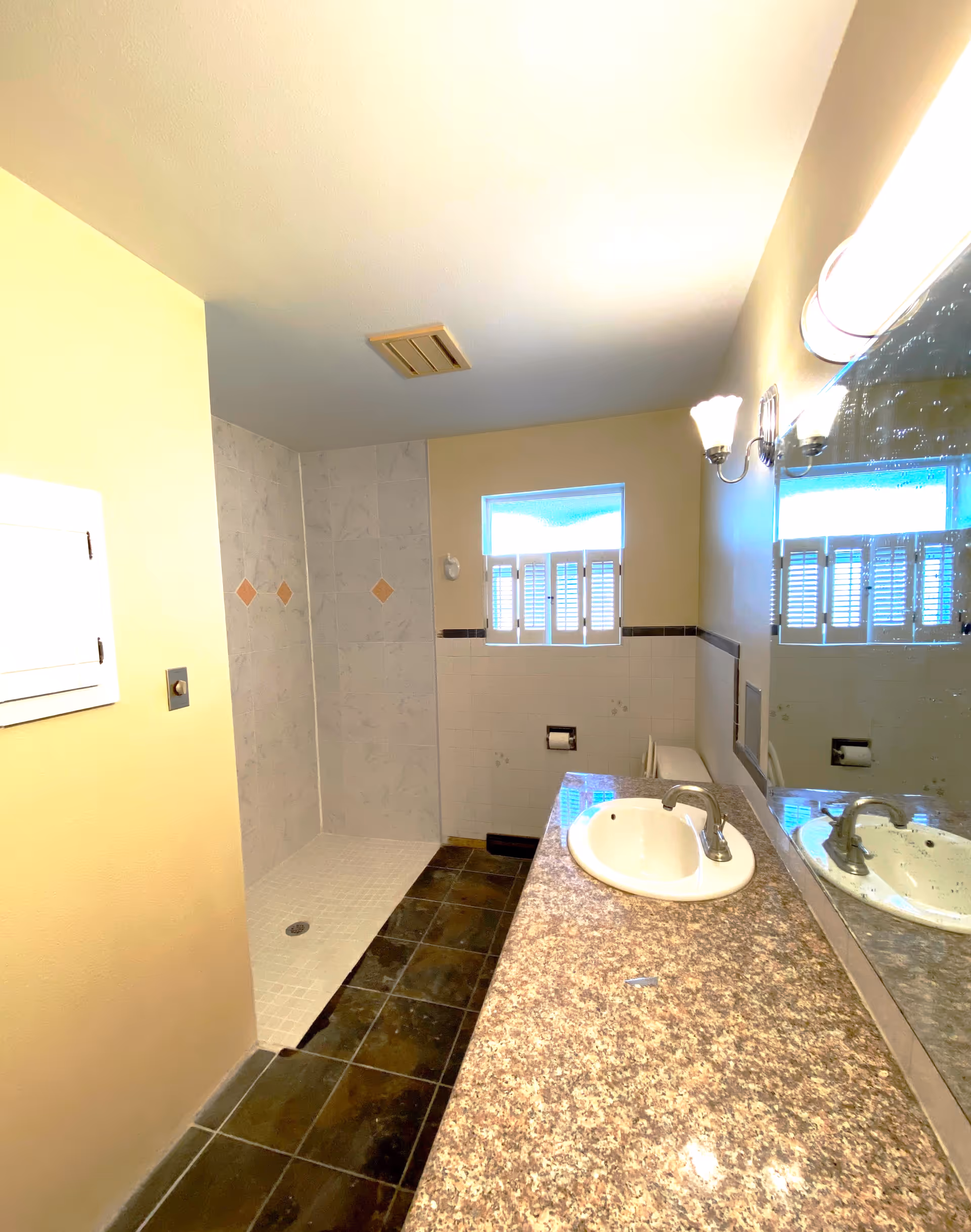 A bright bathroom with double sink, granite countertop, tiled shower, and window blinds.