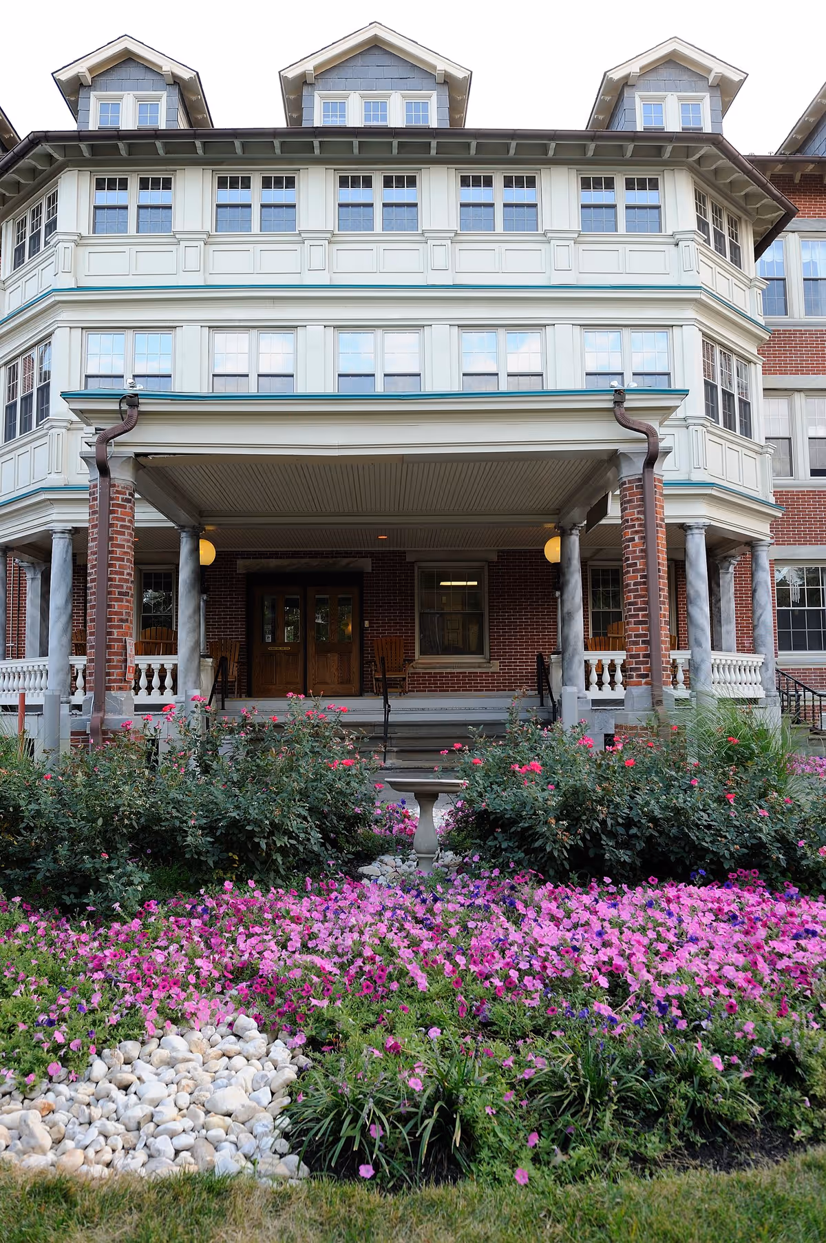 Front exterior view of a multi-story brick building with white trim and multiple windows. The entrance features a covered porch supported by columns, with wooden double doors and rocking chairs on the porch. In front of the building is a garden with pink and purple flowers and a small stone pathway.