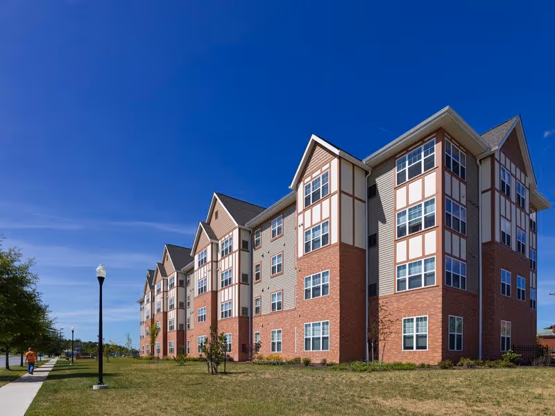 Multi-story brick-and-siding residential building with many windows, a lawn, and a sidewalk under a clear blue sky.