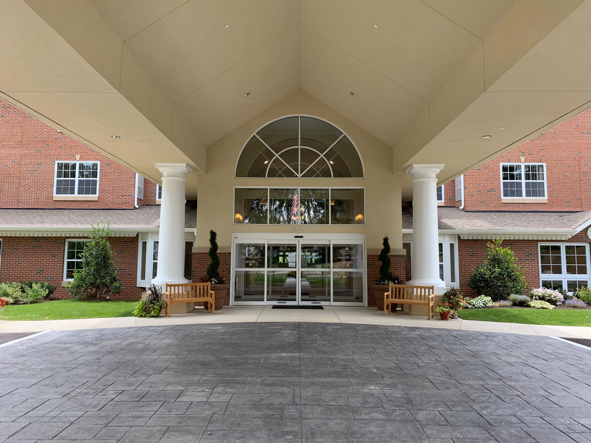 Entrance of a brick building with a covered driveway supported by large white columns. There are two wooden benches and potted plants on either side of the glass double doors. The building has multiple windows and a large arched window above the entrance.