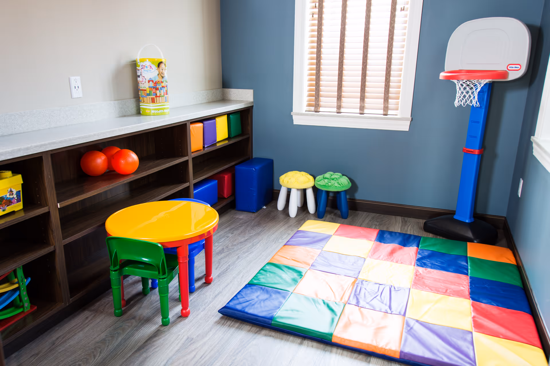 Colorful children's playroom with mat, small table, chairs, shelving, and basketball hoop.