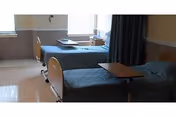 Two hospital-style beds with teal covers, a nightstand, and a window in a shared patient room.
