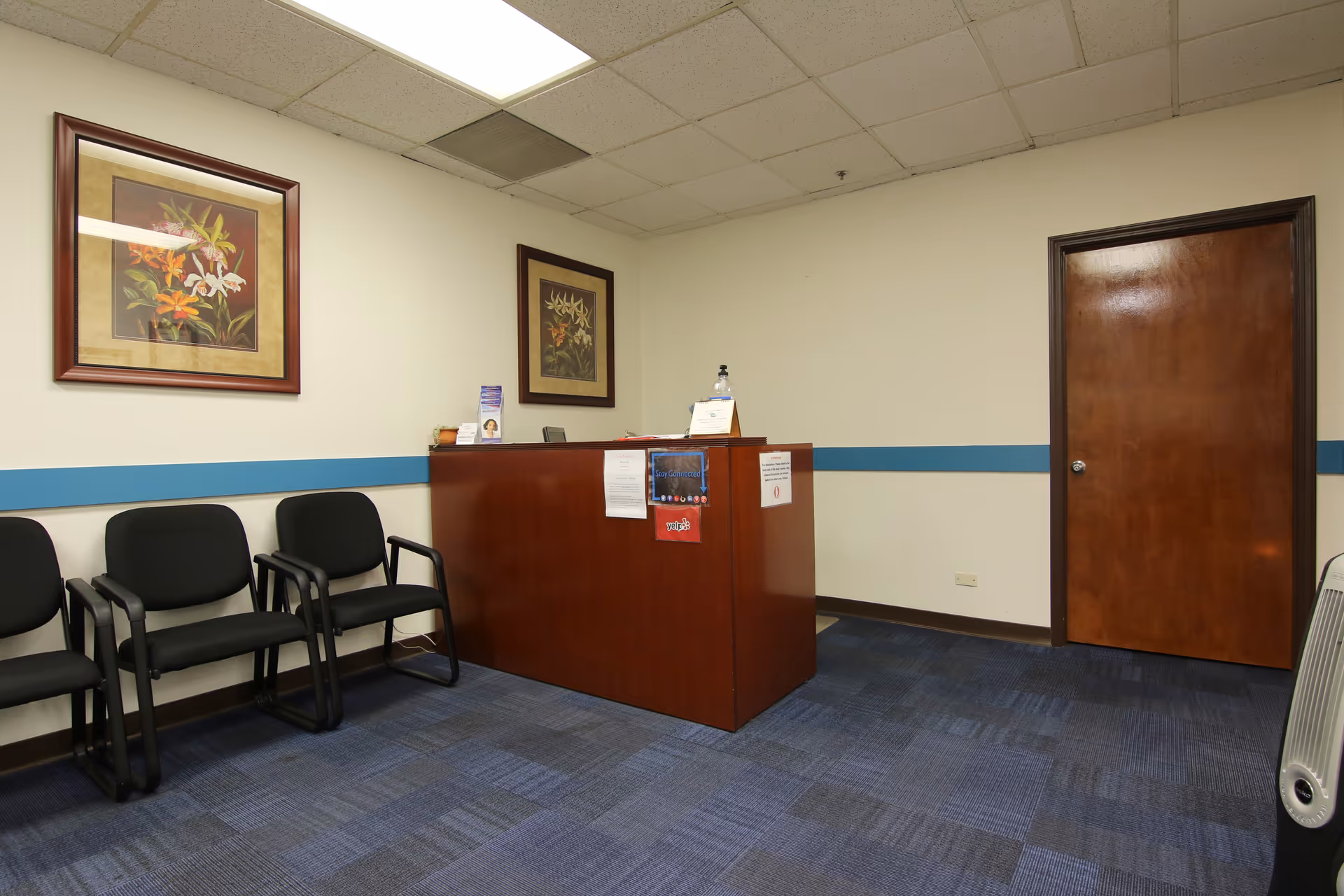 Reception area with a wooden desk, black chairs, framed art, and a blue wall stripe.