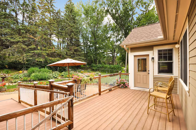 A spacious wooden deck attached to a house with a door and window. The deck has a high chair and railing, and overlooks a garden area with green trees, plants, flowers, and a patio umbrella providing shade over a table and chairs.