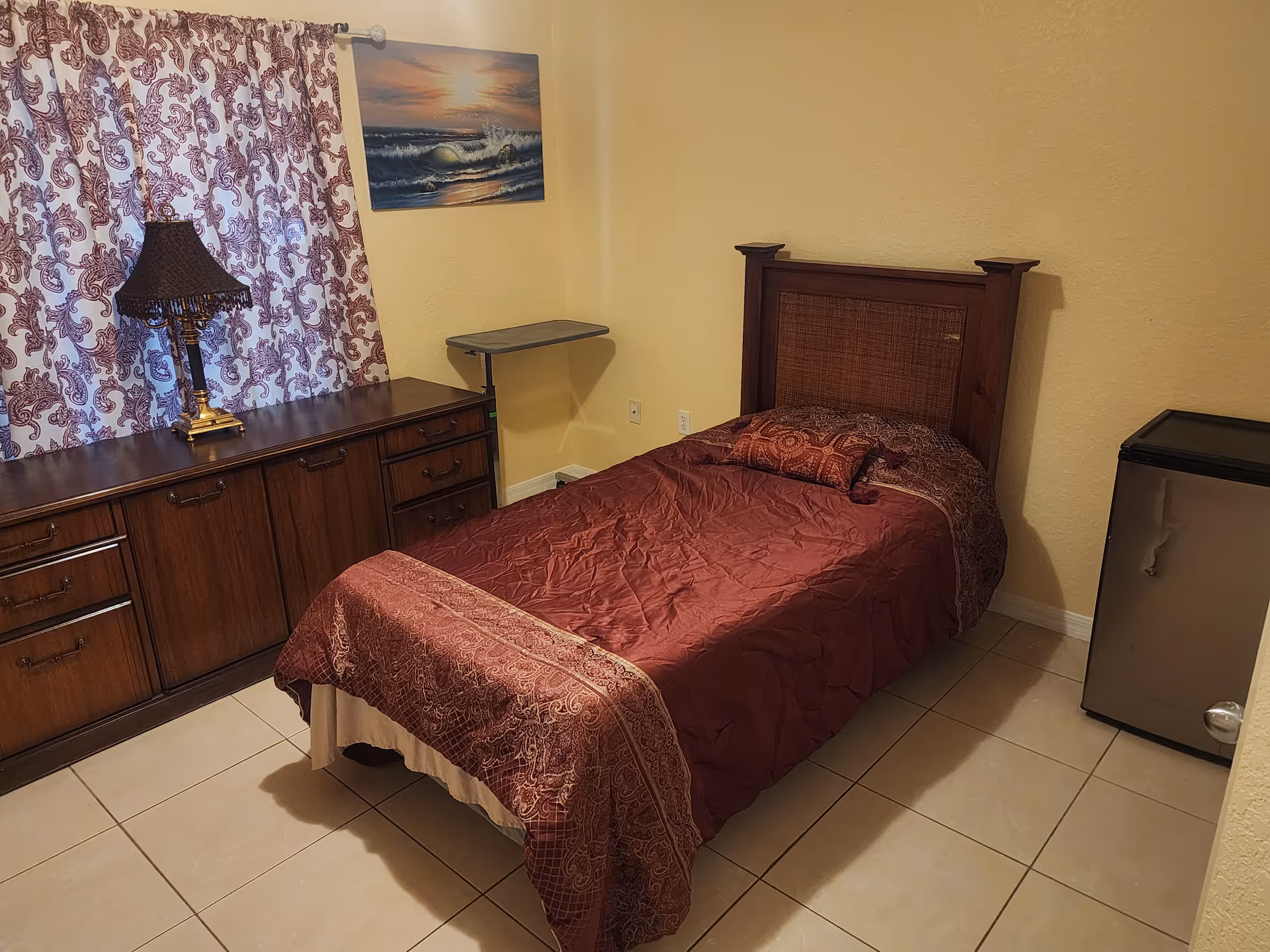 Single bedroom with burgundy bed, dark wood dresser, lamp, curtains, and mini fridge.