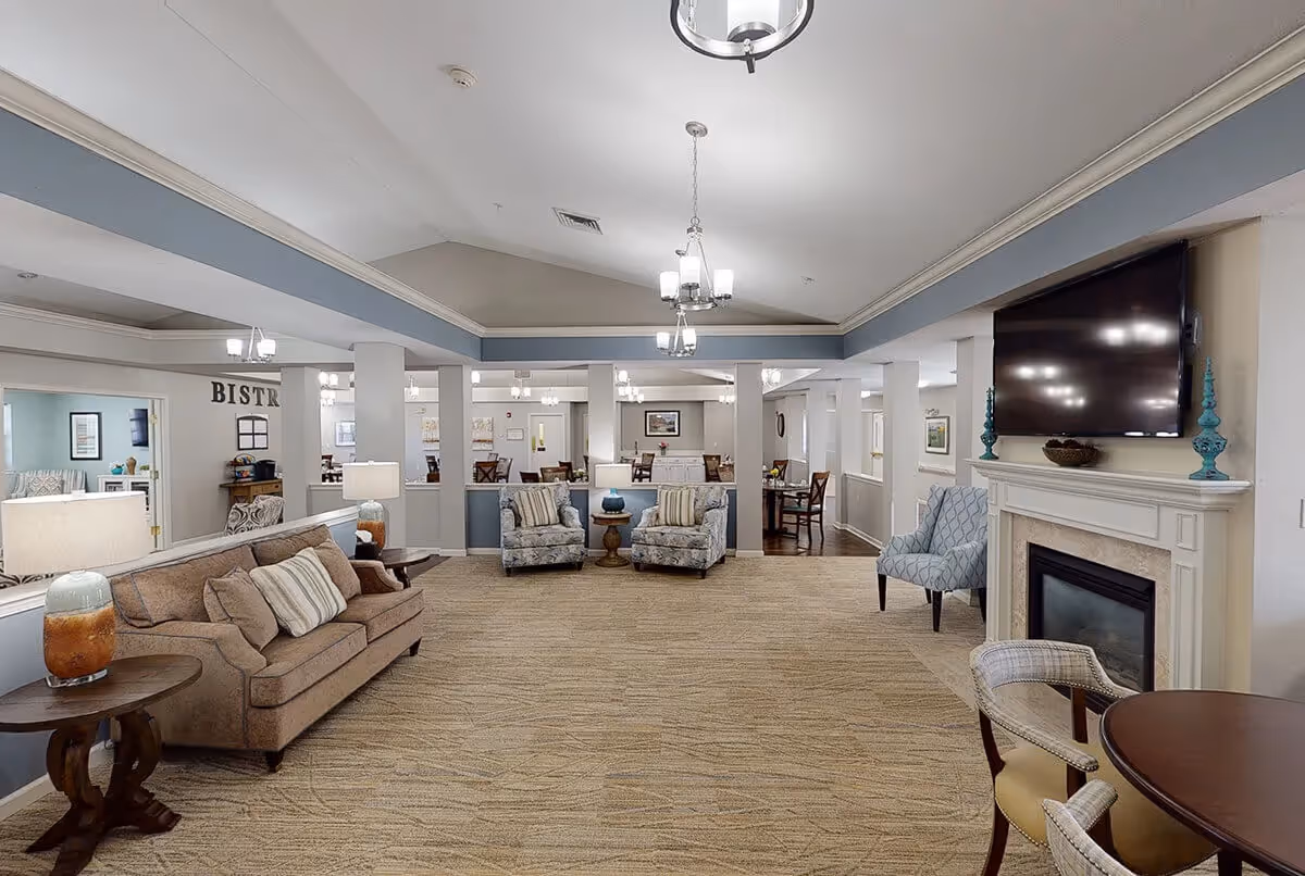A spacious and well-lit living room area with beige carpet, a brown sofa with striped pillows, two patterned armchairs, a fireplace with a mounted flat-screen TV above it, and a round table with chairs. The room has a high ceiling with hanging light fixtures and an open view into a dining area labeled 'Bistro'.