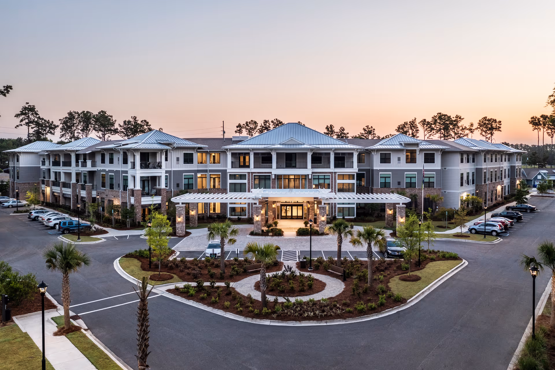 Exterior view of a large, modern three-story senior living facility named The Artisan Mount Pleasant at dusk, with well-maintained landscaping, palm trees, a circular driveway, and parked cars.
