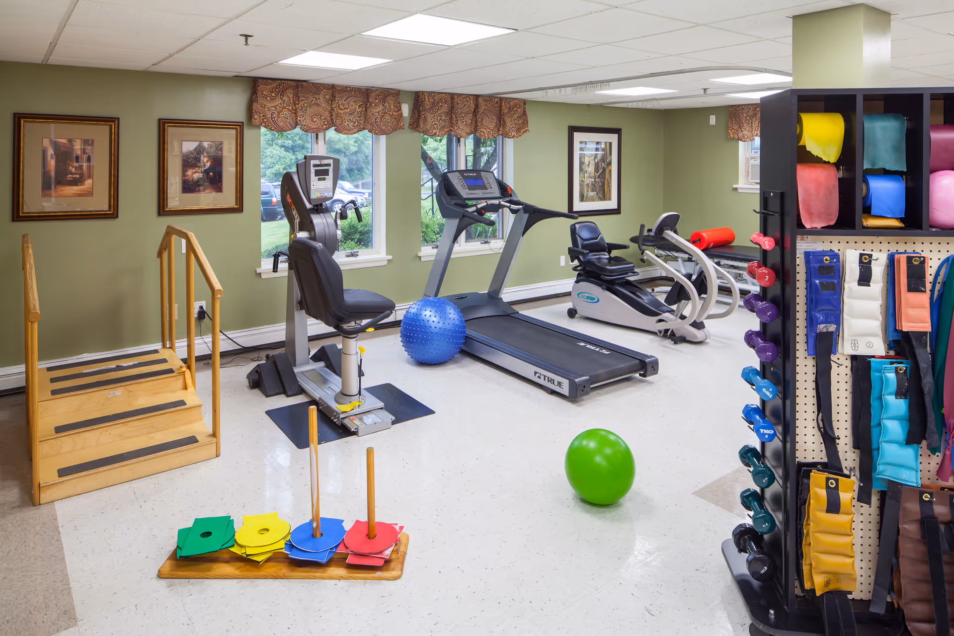 A well-lit exercise room with various fitness equipment including a treadmill, stationary recumbent bike, elliptical machine, colorful exercise balls, hand weights, resistance bands, and a wooden step platform with handrails. The room has light green walls, framed pictures, windows with patterned valances, and a rack holding different colored exercise accessories.