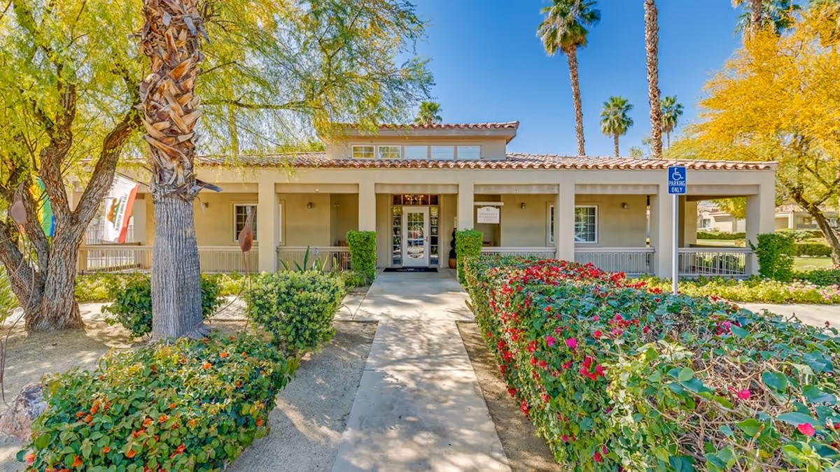 Beige senior living facility entrance framed by palm trees and flowering hedges.