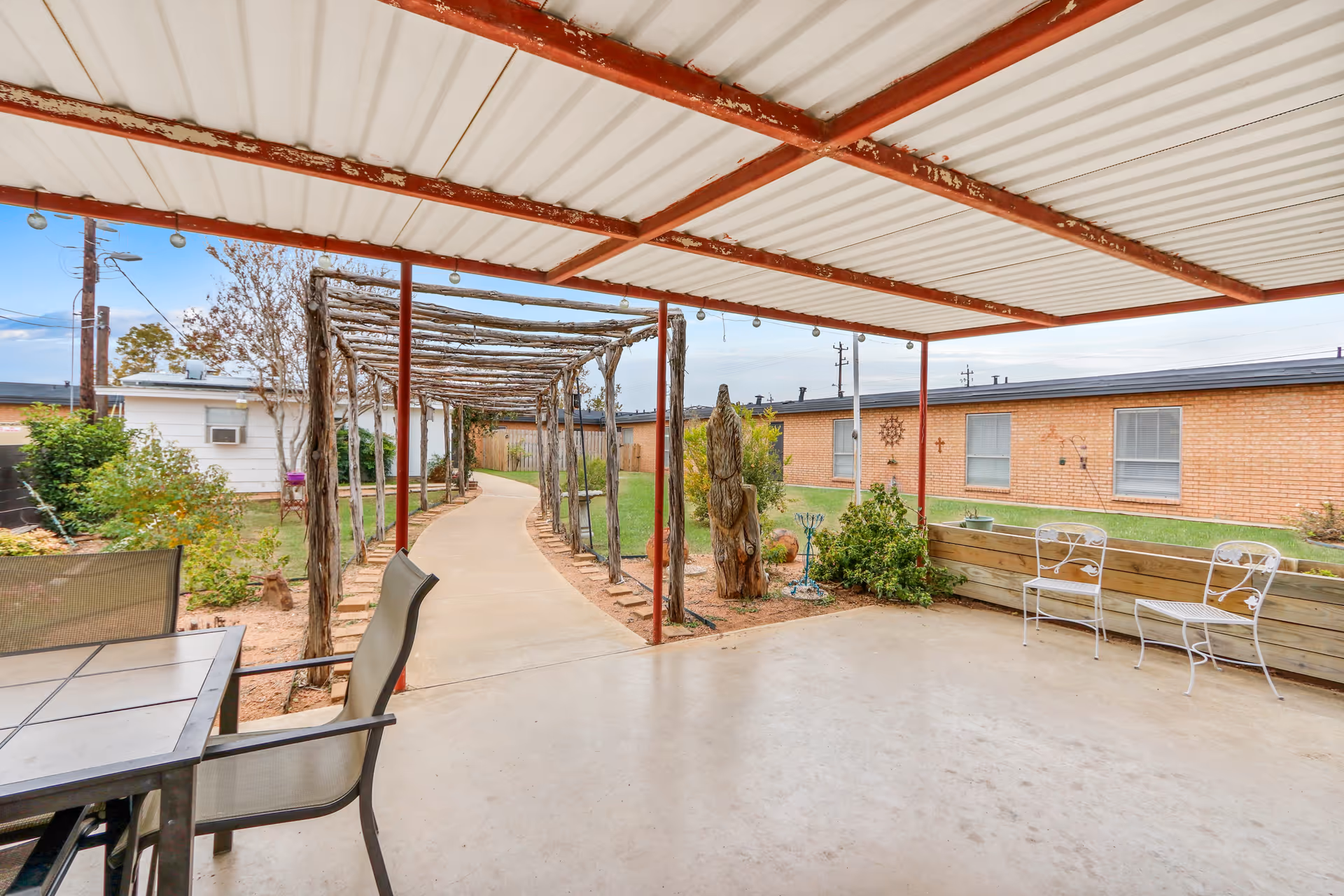 Covered outdoor patio area with a metal roof and a tiled table with chairs on the left. A concrete pathway leads through a wooden pergola structure surrounded by plants and greenery. In the background, there are single-story brick buildings with windows and some outdoor decorations on the walls.