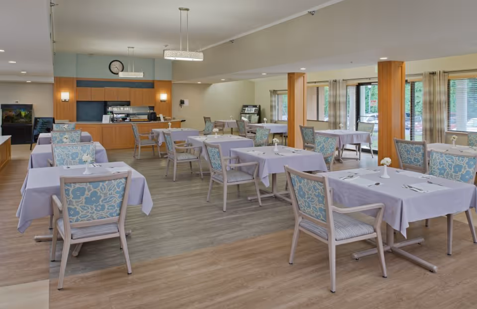 A spacious dining room with multiple tables covered in light purple tablecloths, each set with a small white vase holding a flower and silverware. The chairs have light wood frames with blue and white floral patterned cushions. The room has large windows with beige curtains allowing natural light to enter. At the far end, there is a counter area with a clock above it and some kitchen equipment. The floor is a combination of light wood and vinyl.