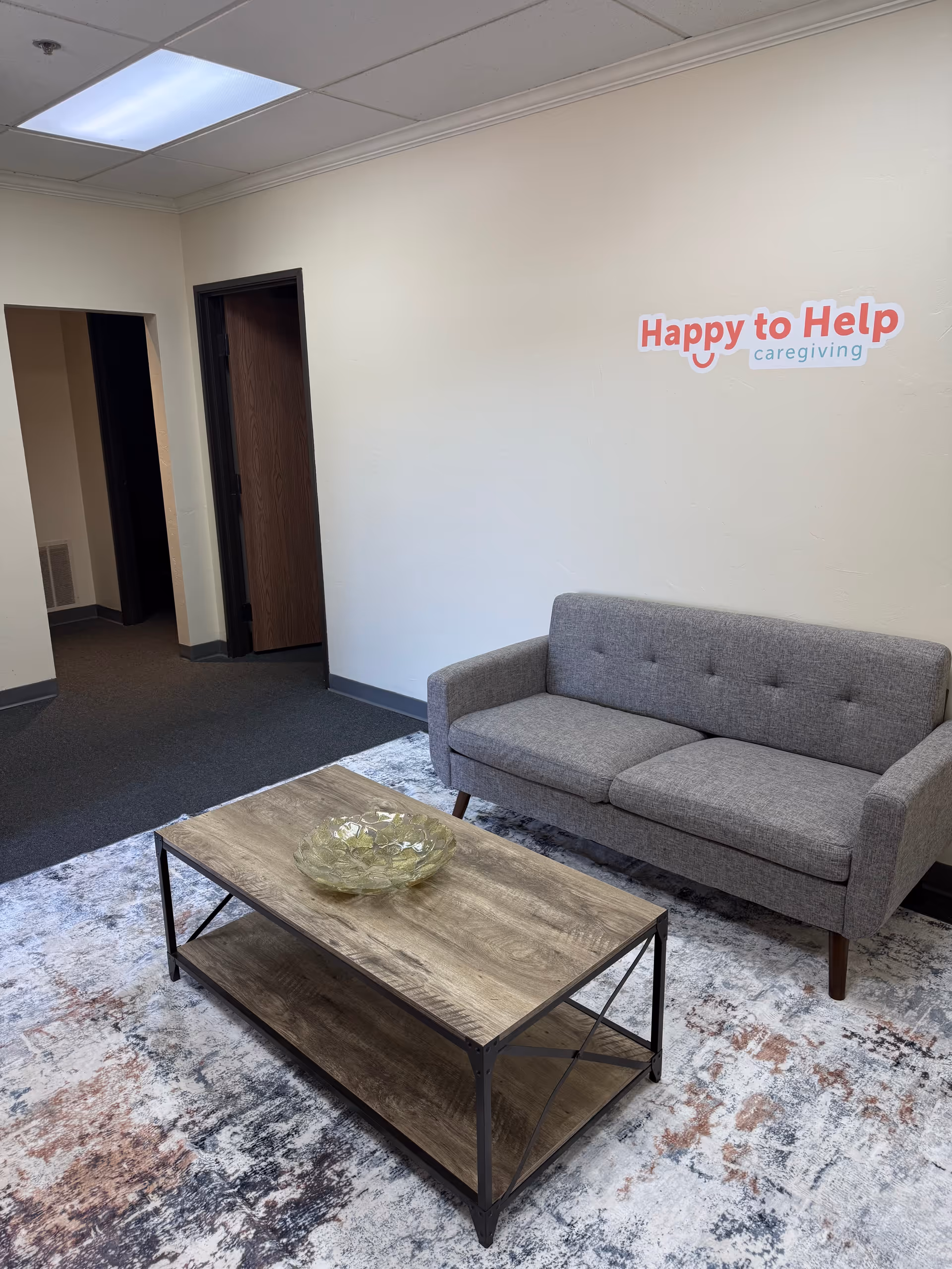 Interior waiting area with gray sofa, coffee table, rug, and 'Happy to Help Caregiving' logo
