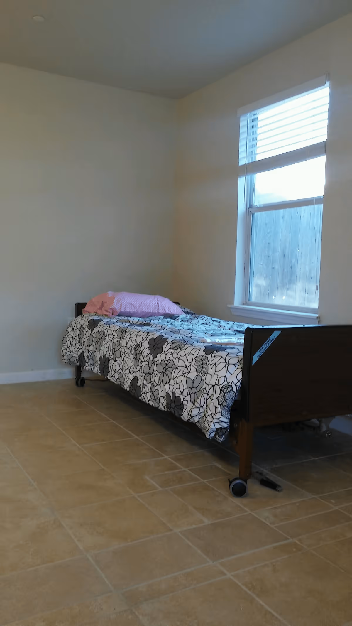 A simple bedroom with a single bed against the wall, covered with a floral patterned bedspread and a pink pillow. There is a window with blinds letting in natural light, and the floor is tiled.