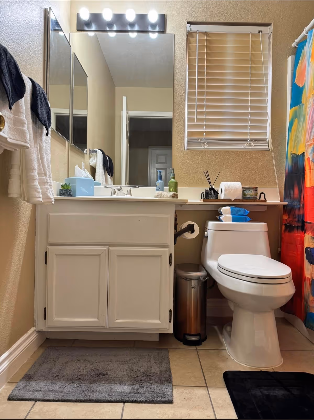 A compact bathroom featuring a white vanity, mirror, toilet, window blind, and shower curtain.