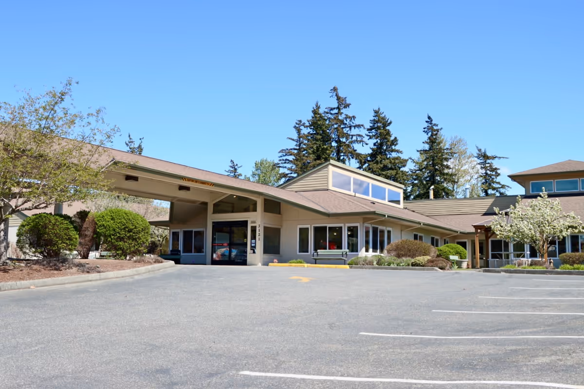 Front entrance of a low-rise senior living facility with a covered drop-off canopy, parking lot, and landscaped shrubs under a clear blue sky.