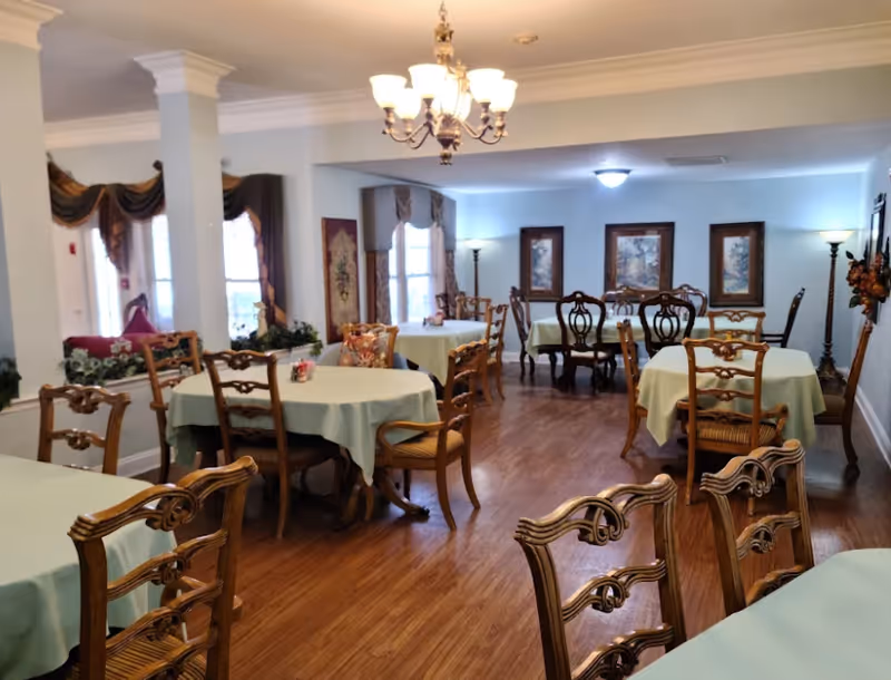 Elegant dining room with round tables, green tablecloths, and wooden chairs in a senior facility.