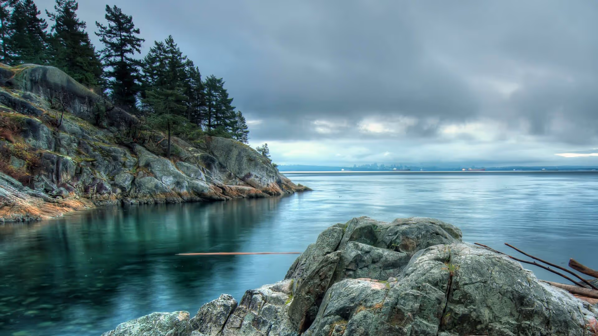 Rocky shoreline with calm water, evergreen trees, and a cloudy sky.