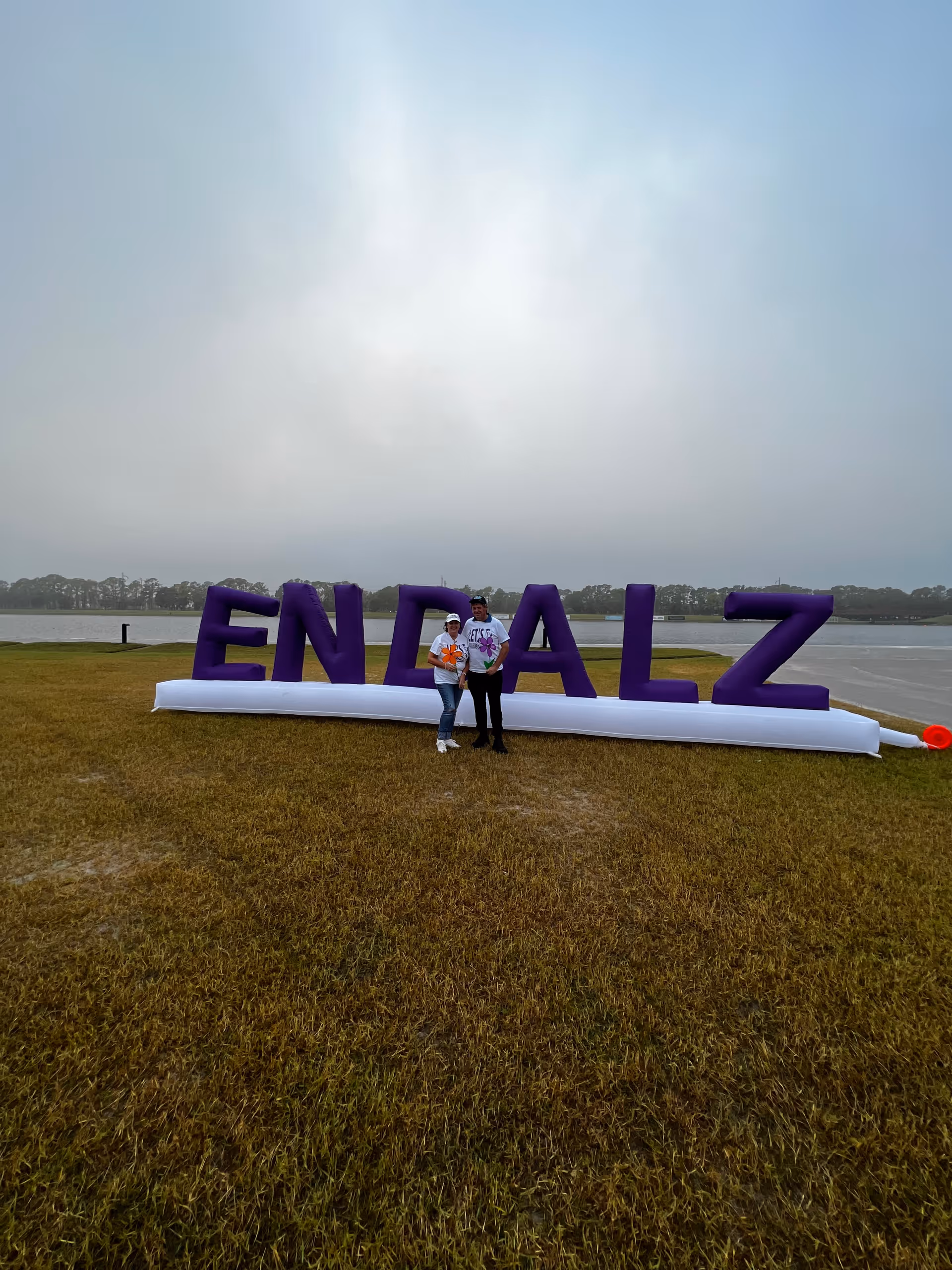 Two people stand in front of large purple ENDALZ letters on a grassy lakeside field.