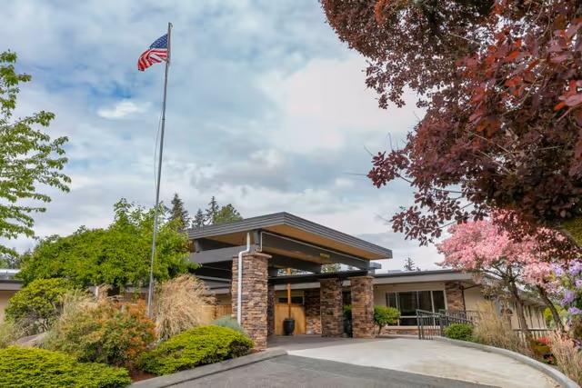 Front entrance of a rehabilitation center with a covered porte-cochere, an American flag, and landscaped shrubs and trees.