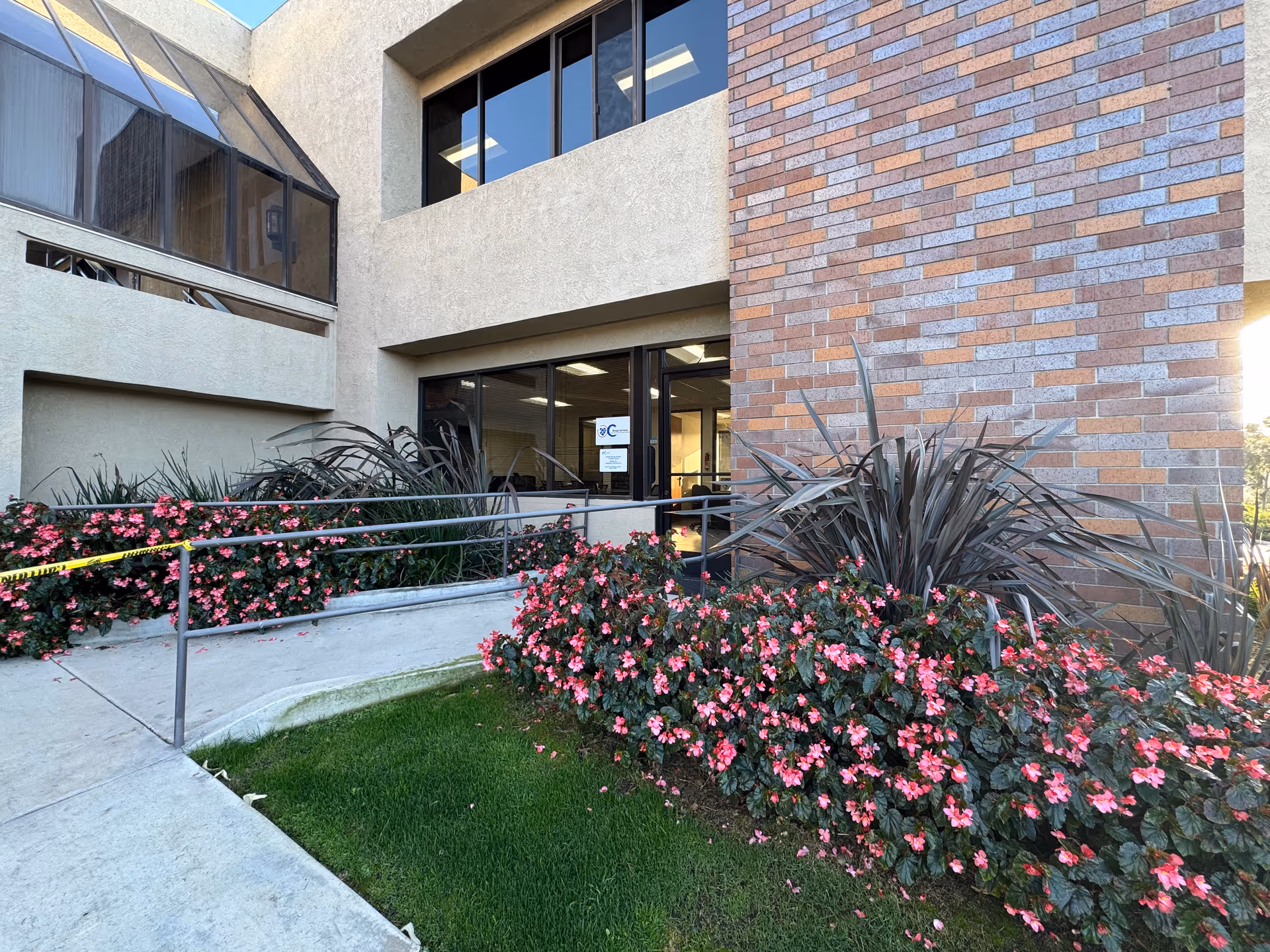 Exterior view of a senior living facility entrance with brick wall, glass doors, and flowering landscaping.