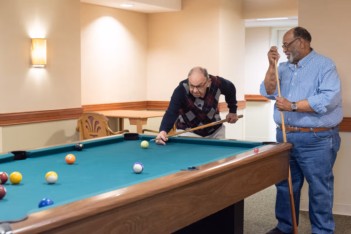 Two elderly men playing pool in a well-lit room with beige walls and wooden trim. One man is leaning over the pool table aiming a shot, while the other stands nearby holding a cue stick and watching.