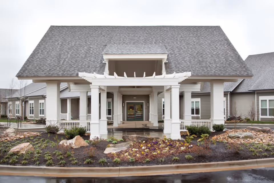 Front exterior view of Concord Place Memory Care facility showing a large covered entrance with white pillars, a gray shingled roof, landscaped garden beds with rocks and plants, and a paved driveway in front.