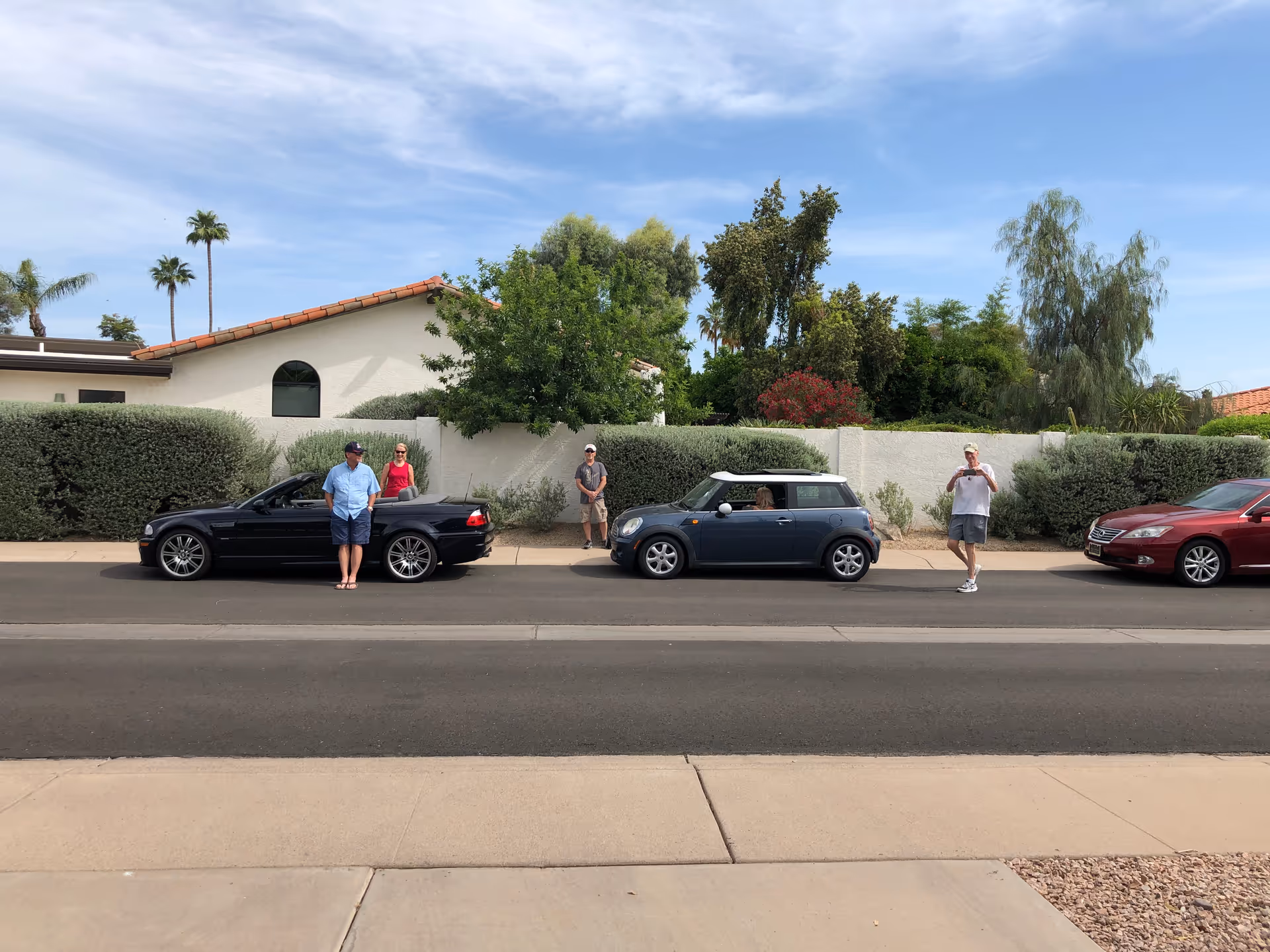 Several people stand beside parked cars along a quiet suburban street in front of a house.