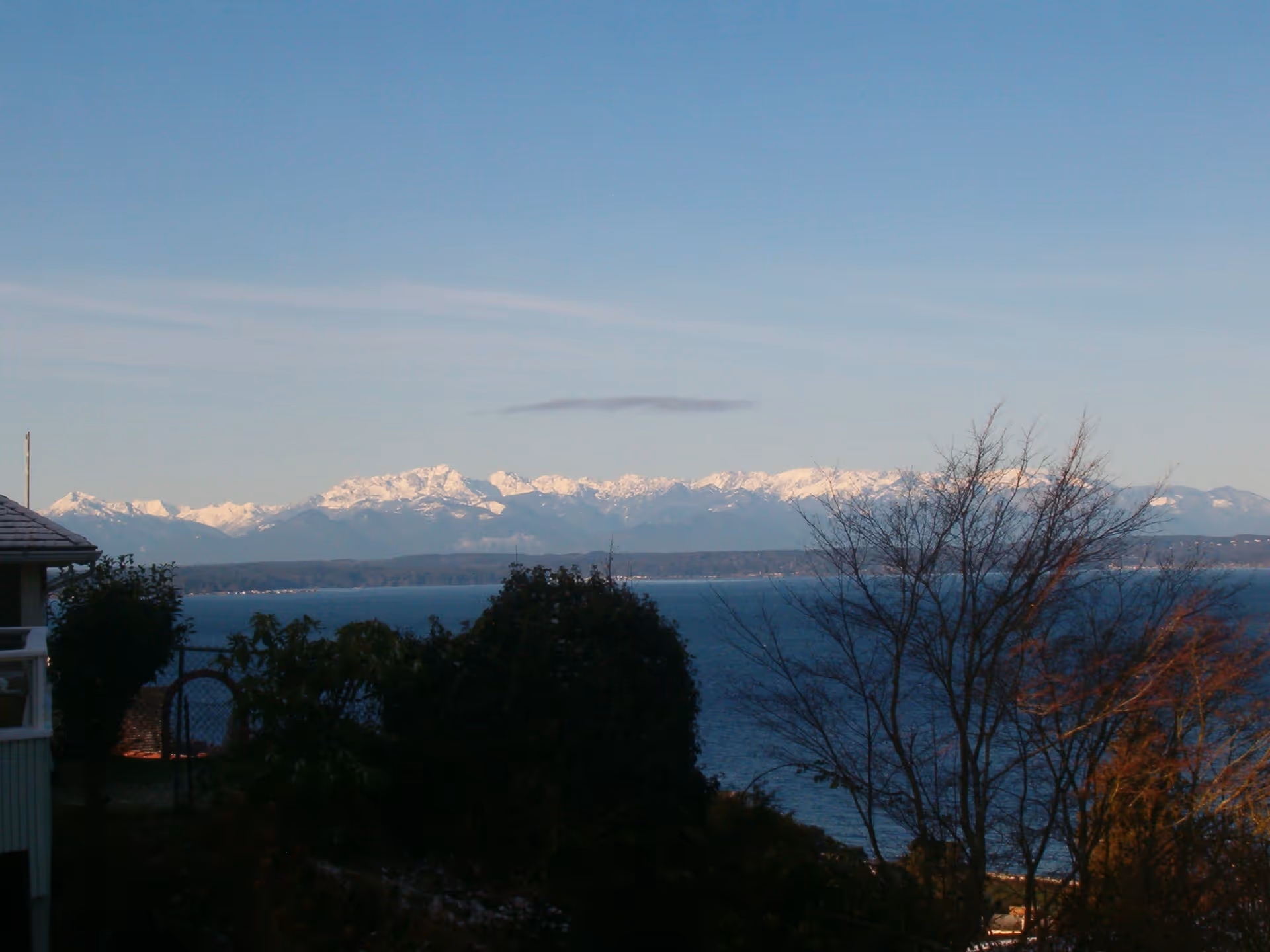 Snowy mountains across a calm lake with leafless trees and a building in foreground.
