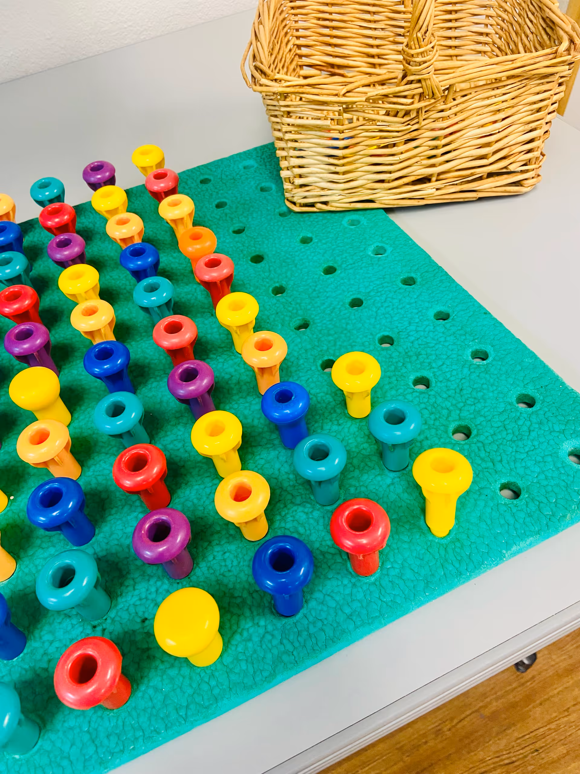 Colorful plastic pegs arranged on a green craft mat beside a wicker basket.