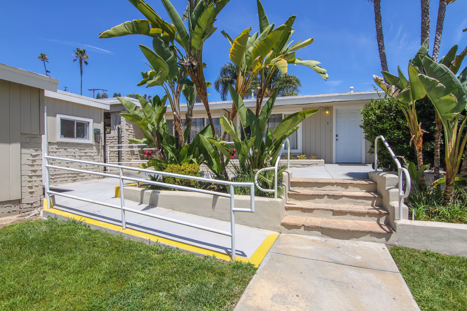 Front exterior of Mesaview Senior Assisted Living with stairs, ramp, and tropical landscaping.