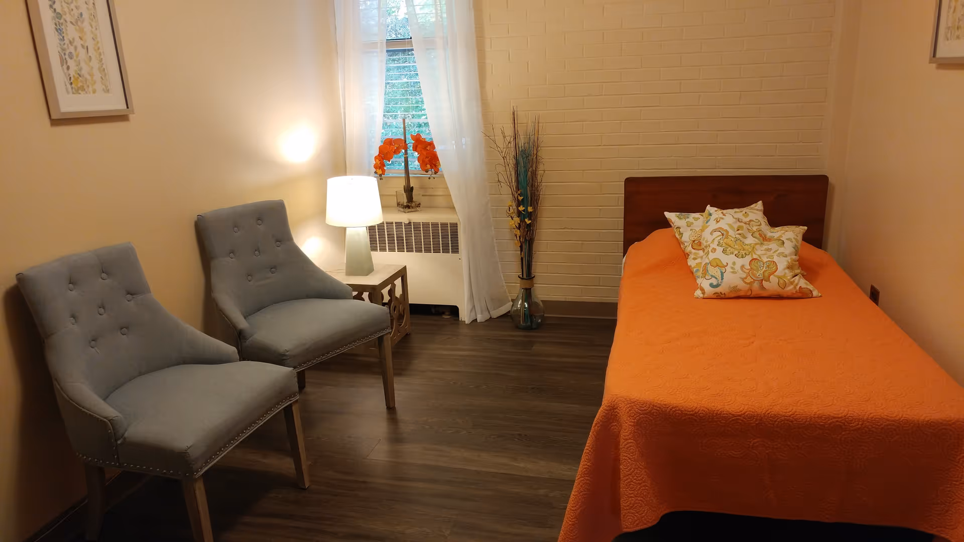 A cozy bedroom with a single bed covered in an orange bedspread and three decorative pillows. Two gray upholstered chairs are placed against the opposite wall. A small table with a lamp and a window with sheer white curtains are between the chairs and the bed. A vase with decorative branches is placed near the bed against a light brick wall.