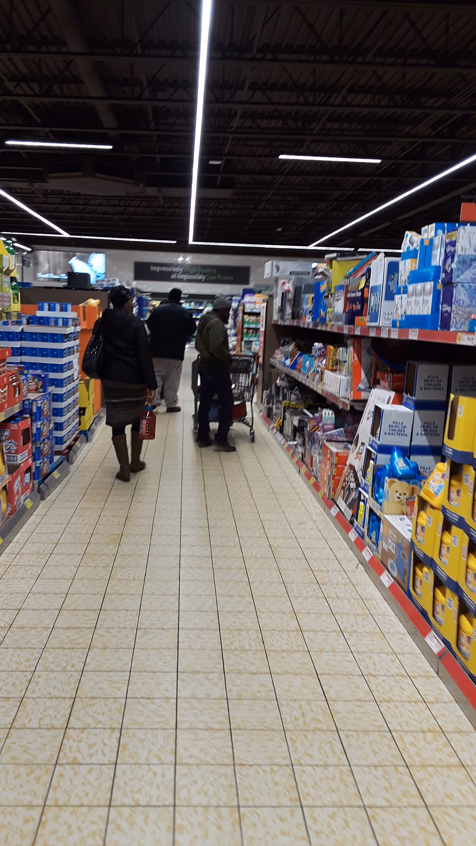 Shoppers move through a brightly lit grocery store aisle with tall shelves.