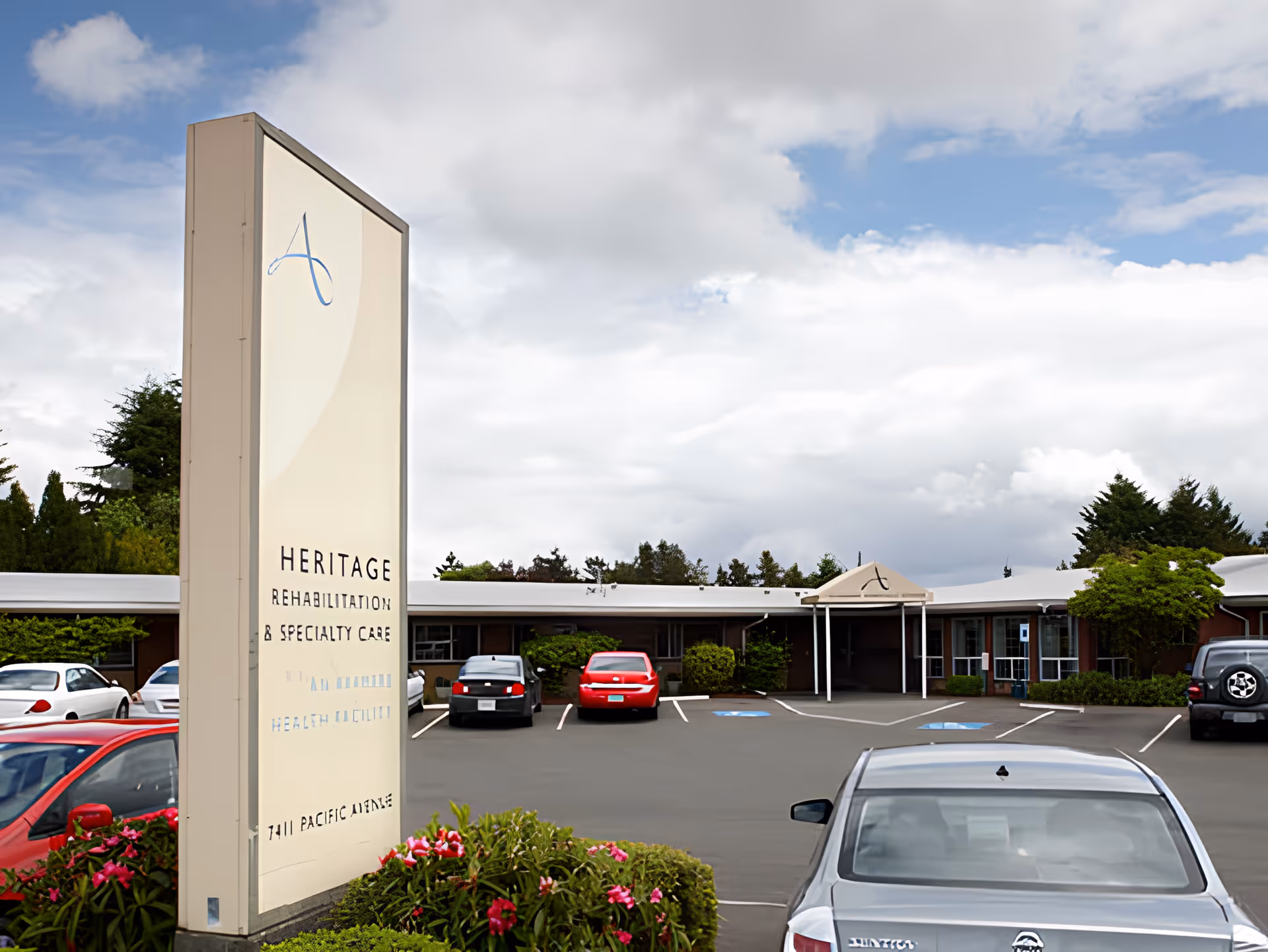 One-story healthcare/rehabilitation building front with a parking lot, cars, and a tall sign reading 'Heritage Rehabilitation & Specialty Care' under a cloudy sky.