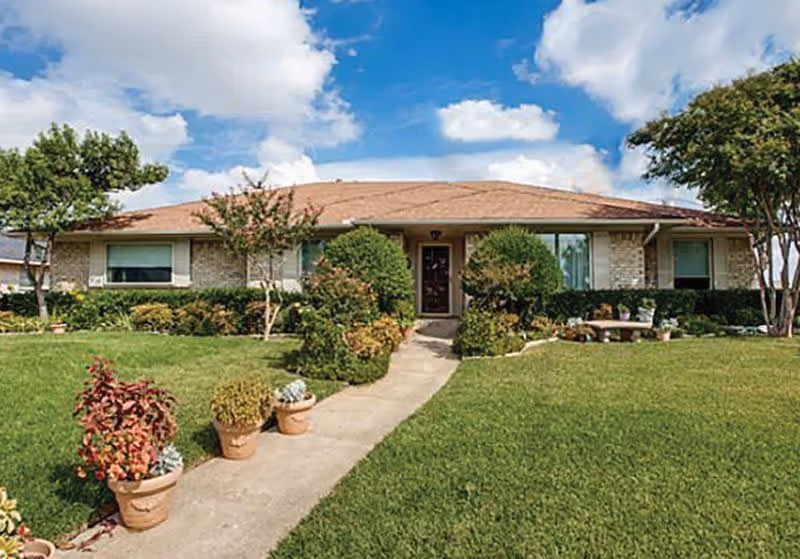 Single-story brick house with a brown roof, surrounded by a well-maintained lawn and various shrubs and potted plants along a concrete walkway leading to the front door under a partly cloudy blue sky.