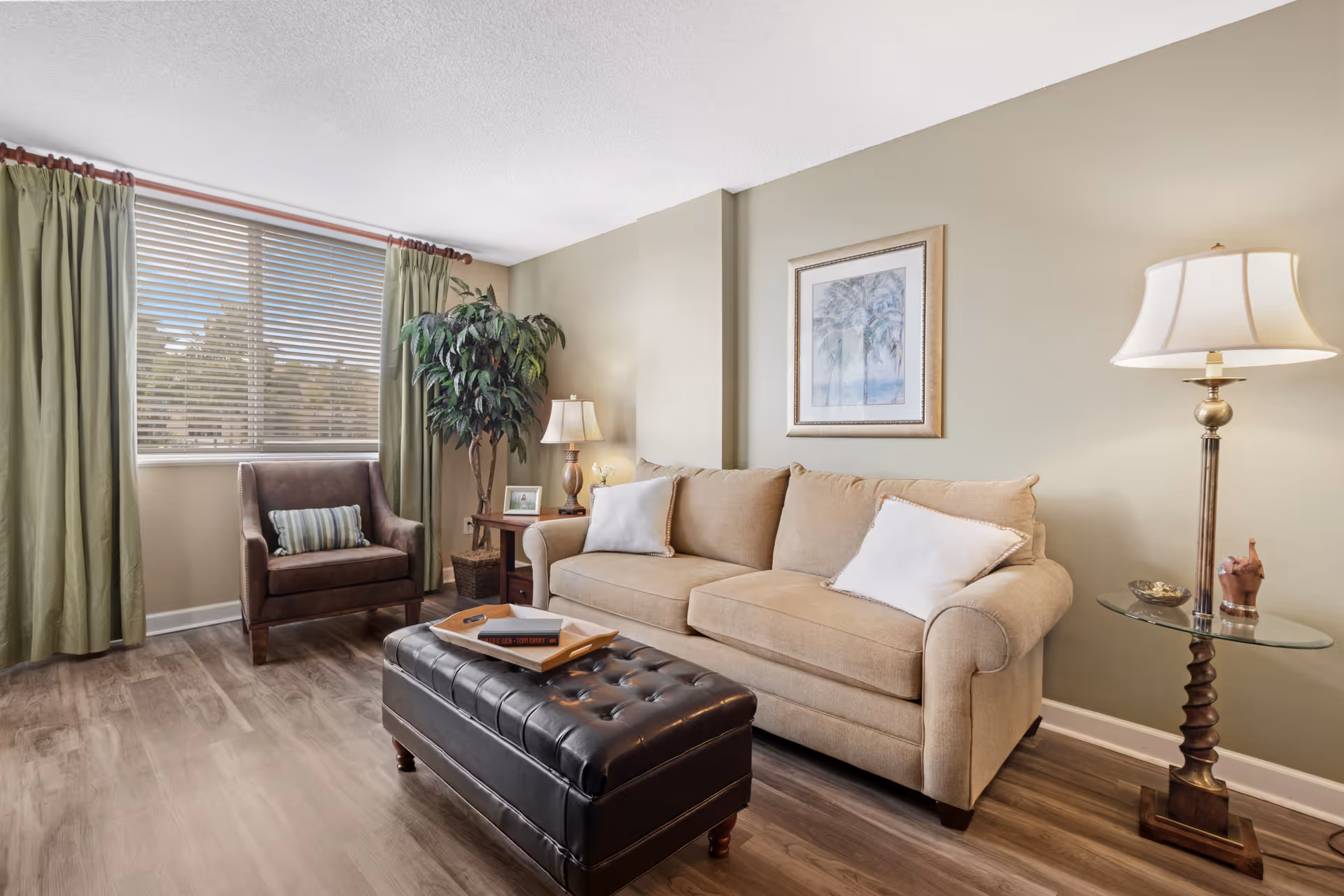 Well-lit living room with a beige sofa, tufted leather ottoman, armchair, floor lamp and a window with green curtains.