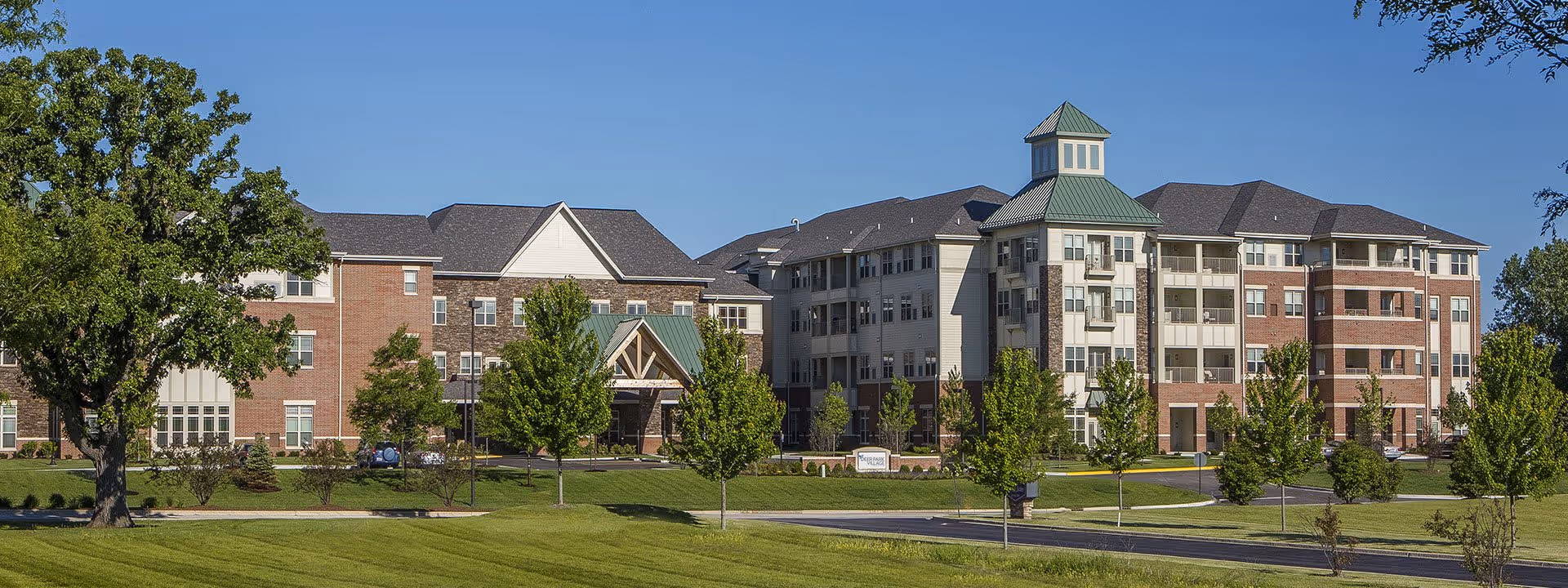 Front view of a multi-story brick and siding senior living building with a cupola, landscaped lawn and trees under a clear blue sky.