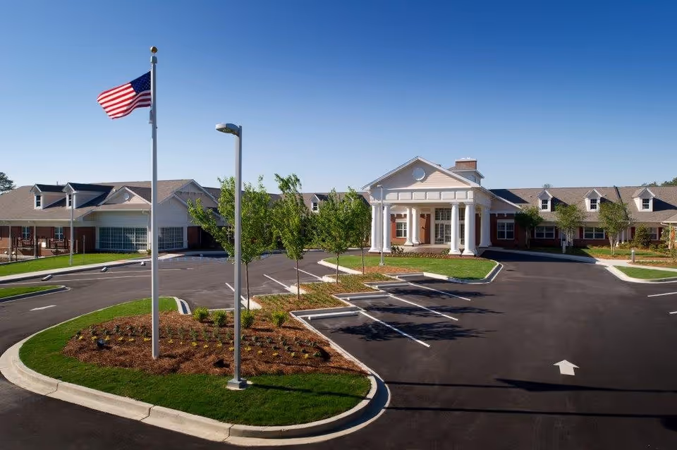 Front entrance of a senior living facility with white columns, an American flag, landscaped islands, and an empty parking lot under a clear blue sky.