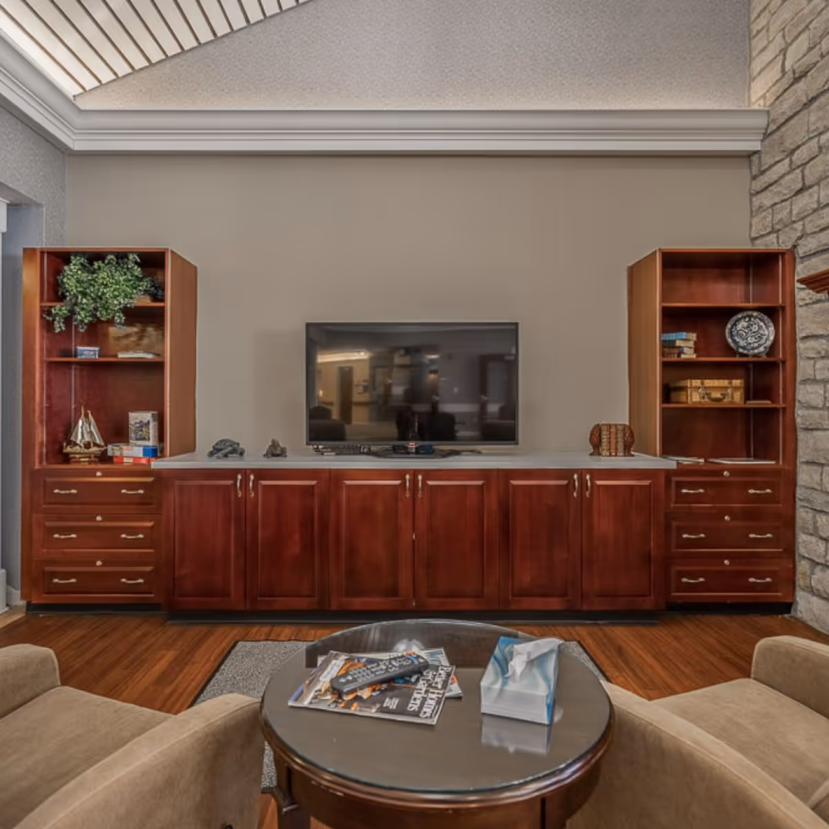 A cozy living room area featuring a flat-screen TV mounted on a wall with wooden cabinets and shelves on either side. The shelves hold decorative items including books, a model sailboat, and a plant. In front of the TV is a round glass-top coffee table with magazines, a remote control, and a box of tissues. Two beige armchairs are partially visible on either side of the table. The room has wooden flooring and a stone accent wall on the right.