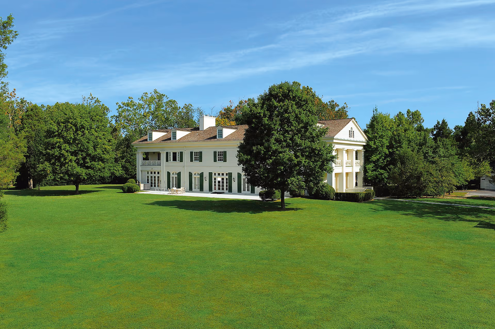 White two-story building with columns sits on a broad green lawn beneath a clear blue sky.