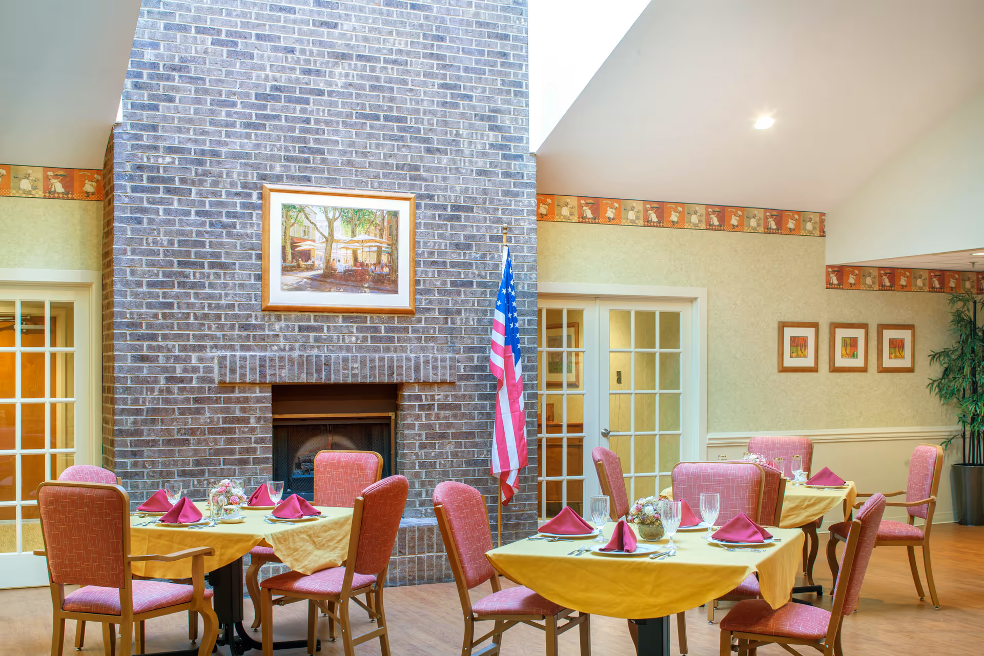 A dining room with several tables covered in yellow tablecloths, each set with pink folded napkins, glassware, and silverware. The room features a large brick fireplace with a framed painting above it and an American flag standing next to it. The walls are decorated with a wallpaper border and framed artwork, and there are glass-paneled double doors in the background.