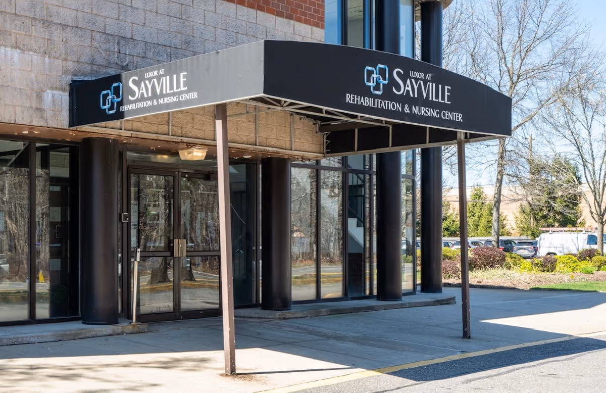 Entrance of Luxor at Sayville Rehabilitation & Nursing Center with a black canopy displaying the facility's name. The building exterior features large glass windows and black pillars, with a sidewalk and some greenery visible nearby.