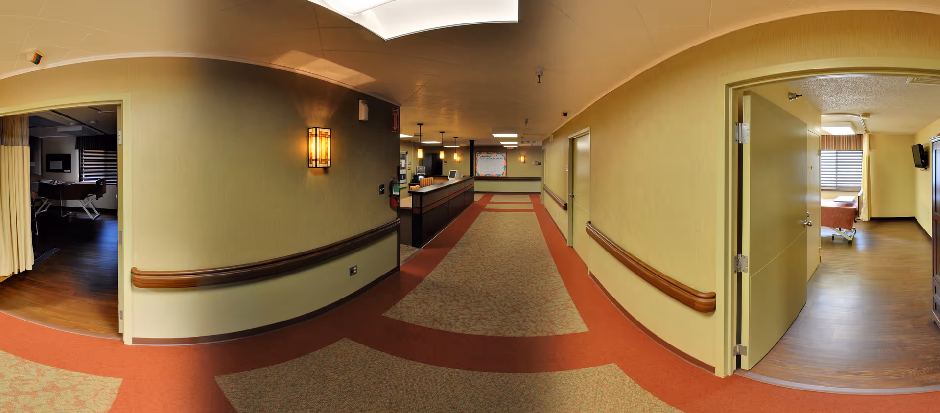 A wide hallway in a senior living facility with beige walls and a patterned carpet. On the left and right sides of the hallway are open doors leading to rooms with beds and windows. The hallway has handrails along the walls and a reception desk visible in the distance under warm lighting.