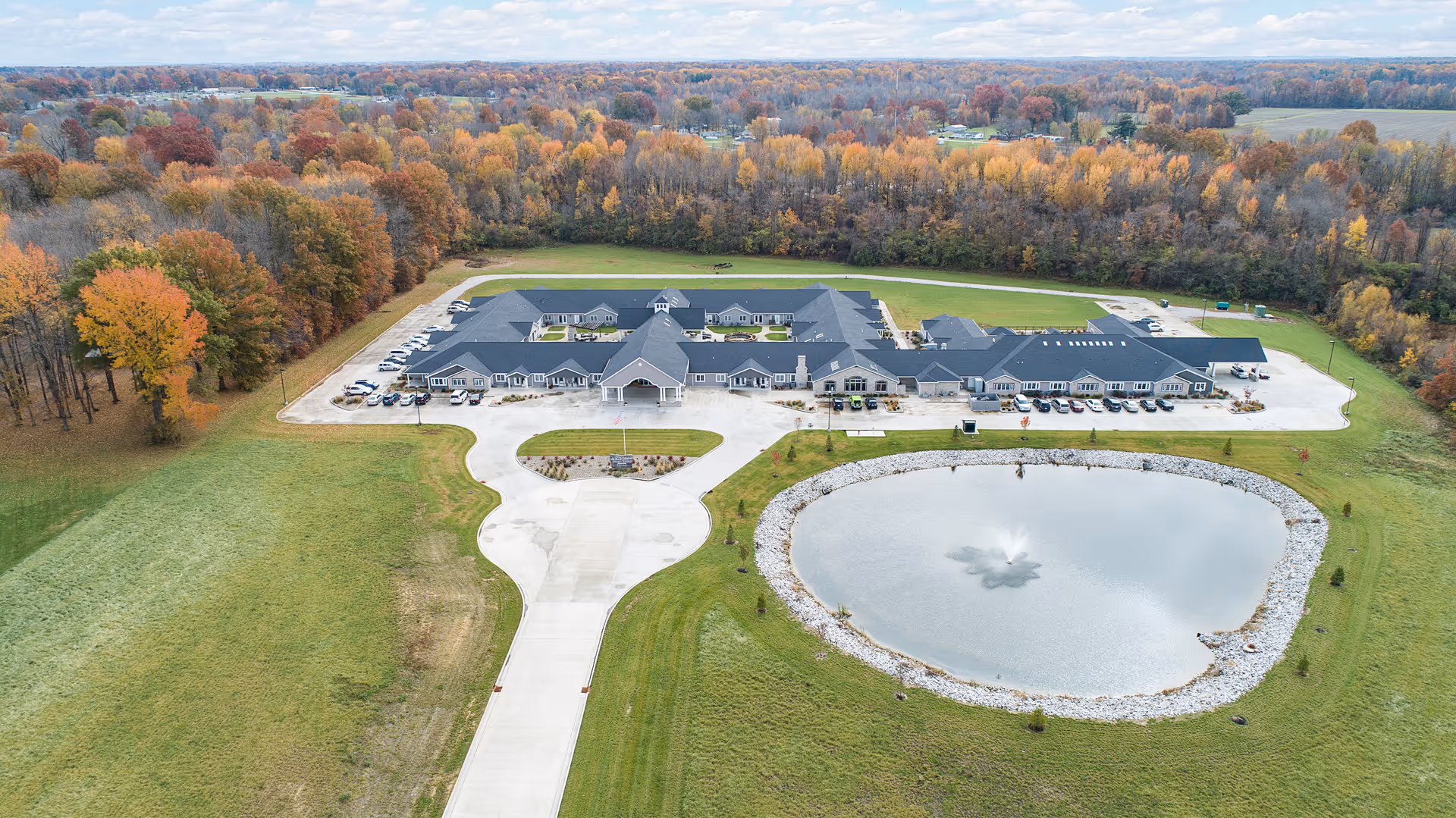 Aerial view of Villas of Holly Brook Assisted Living & Memory Care facility in Brazil, IN, showing a large single-story building with a dark roof surrounded by green lawns and a pond with a fountain in front. The facility is bordered by trees with autumn foliage and a driveway leading to the entrance with parked cars.