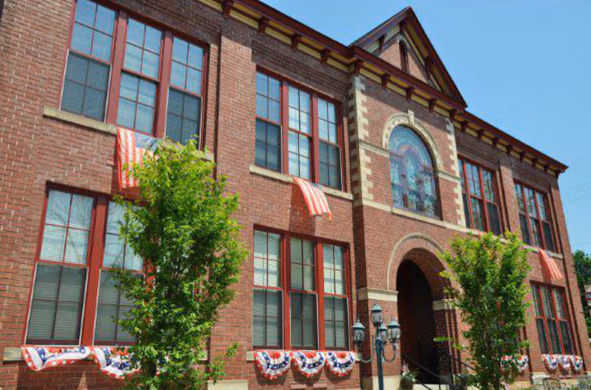 Exterior view of a two-story brick building decorated with American flags and patriotic bunting. The building has large windows with red frames and a central arched entrance. There are small trees and a vintage-style street lamp in front of the building under a clear blue sky.
