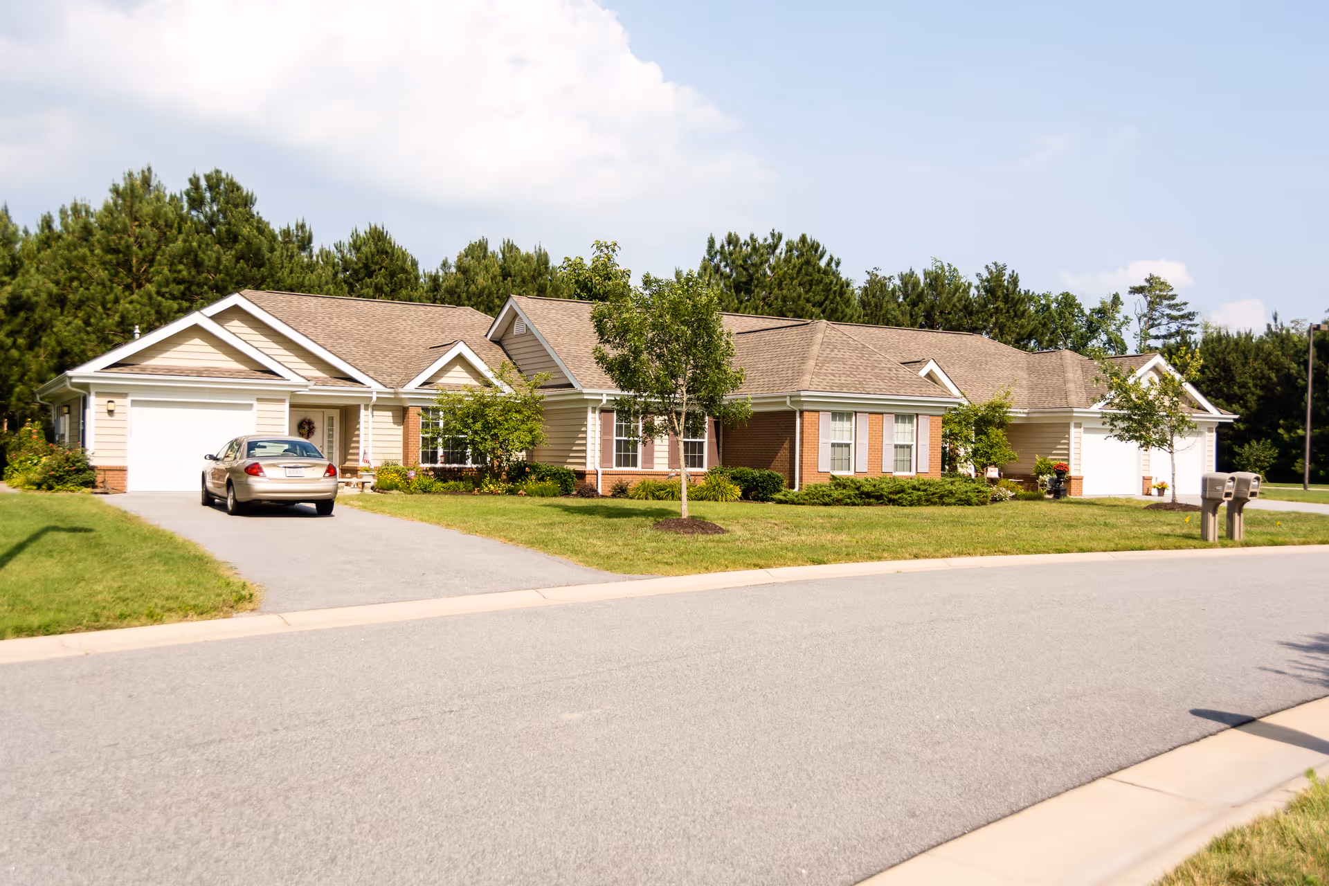 Exterior view of a single-story residential building with beige siding and brick accents, surrounded by green lawns and trees under a partly cloudy sky. A car is parked in the driveway, and there are mailboxes near the street.
