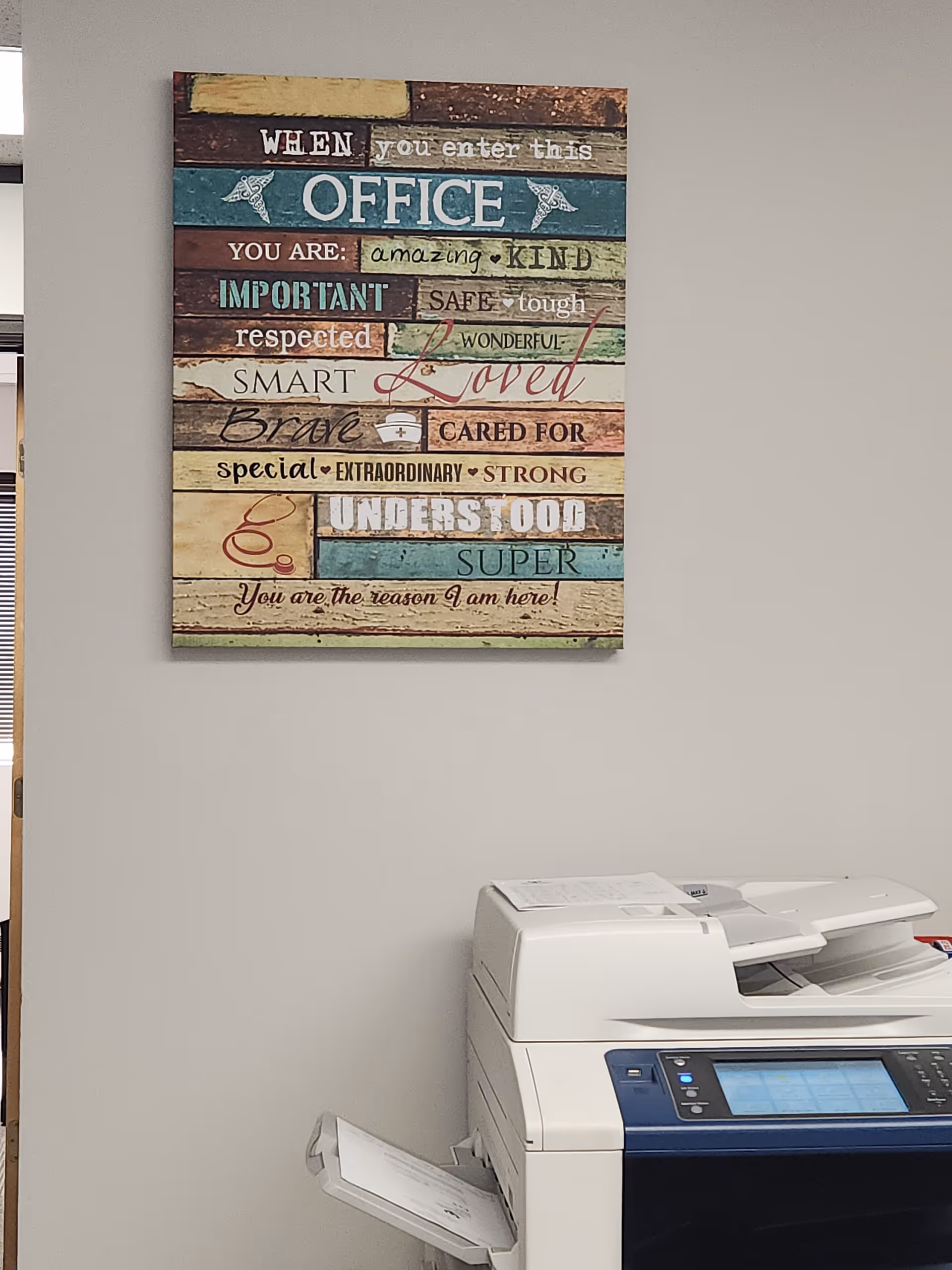 Colorful wooden sign with uplifting words on wall beside a printer in an office.
